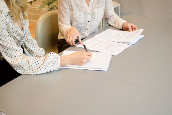 Woman in white shirt with black polka dots signing on white paper with a black pen beside woman in solid white shirts about to pick up documents