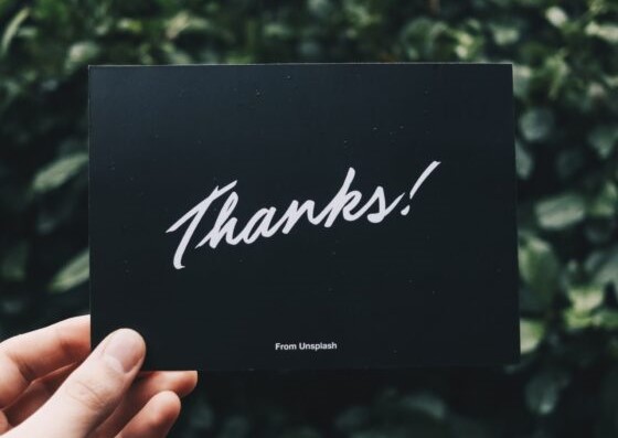A black card with the word “Thanks!” written in white font in the foreground, green leaves or a bush in the background