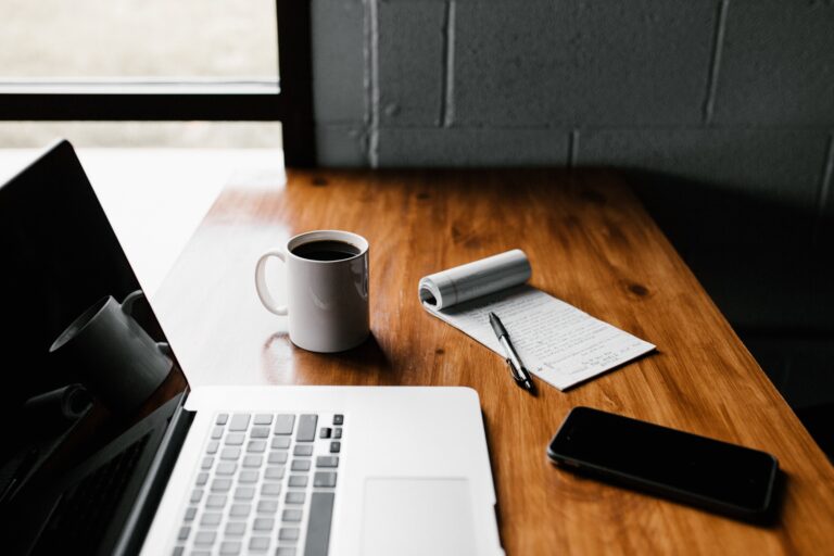 Silver laptop with black screen on wooden table. Cell phone, note pad, and cup of coffee also on table.