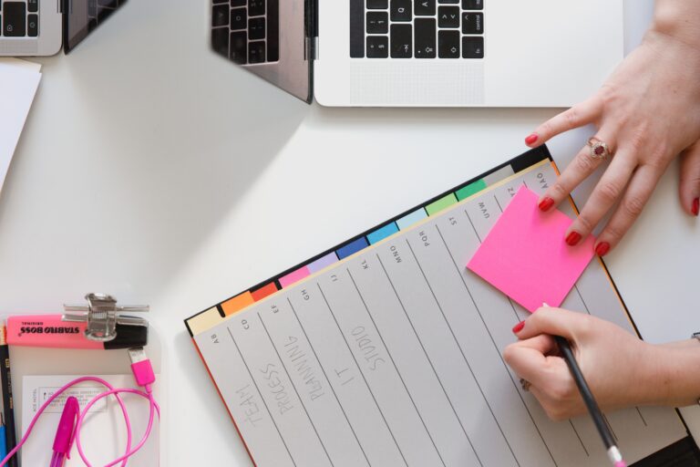 Woman writing on pink post-it note with a daily planner and laptop in the background.